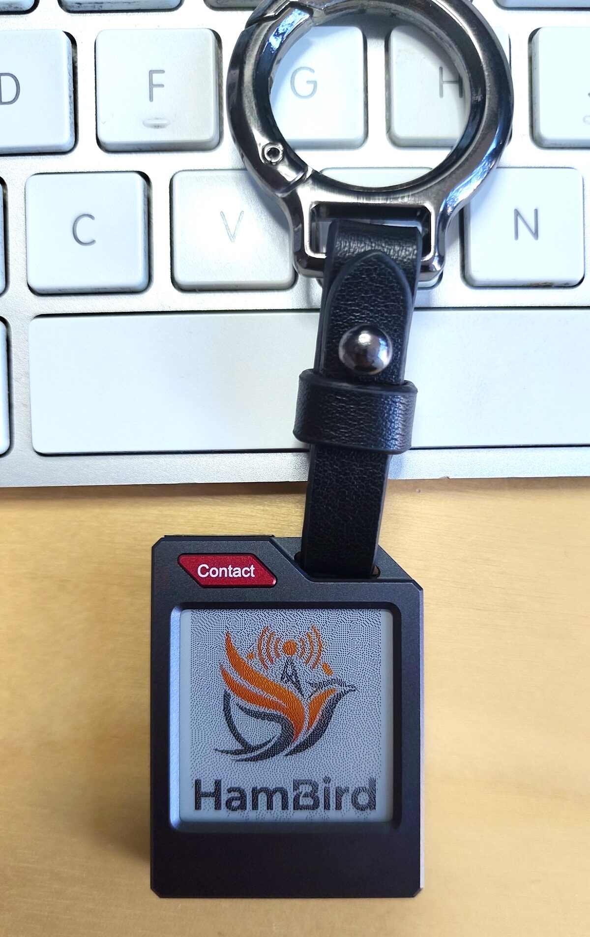 HamBird receiver on a keychain in front of a keyboard: black case, red contact button, 1.54-inch e-ink display showing the HamBird logo